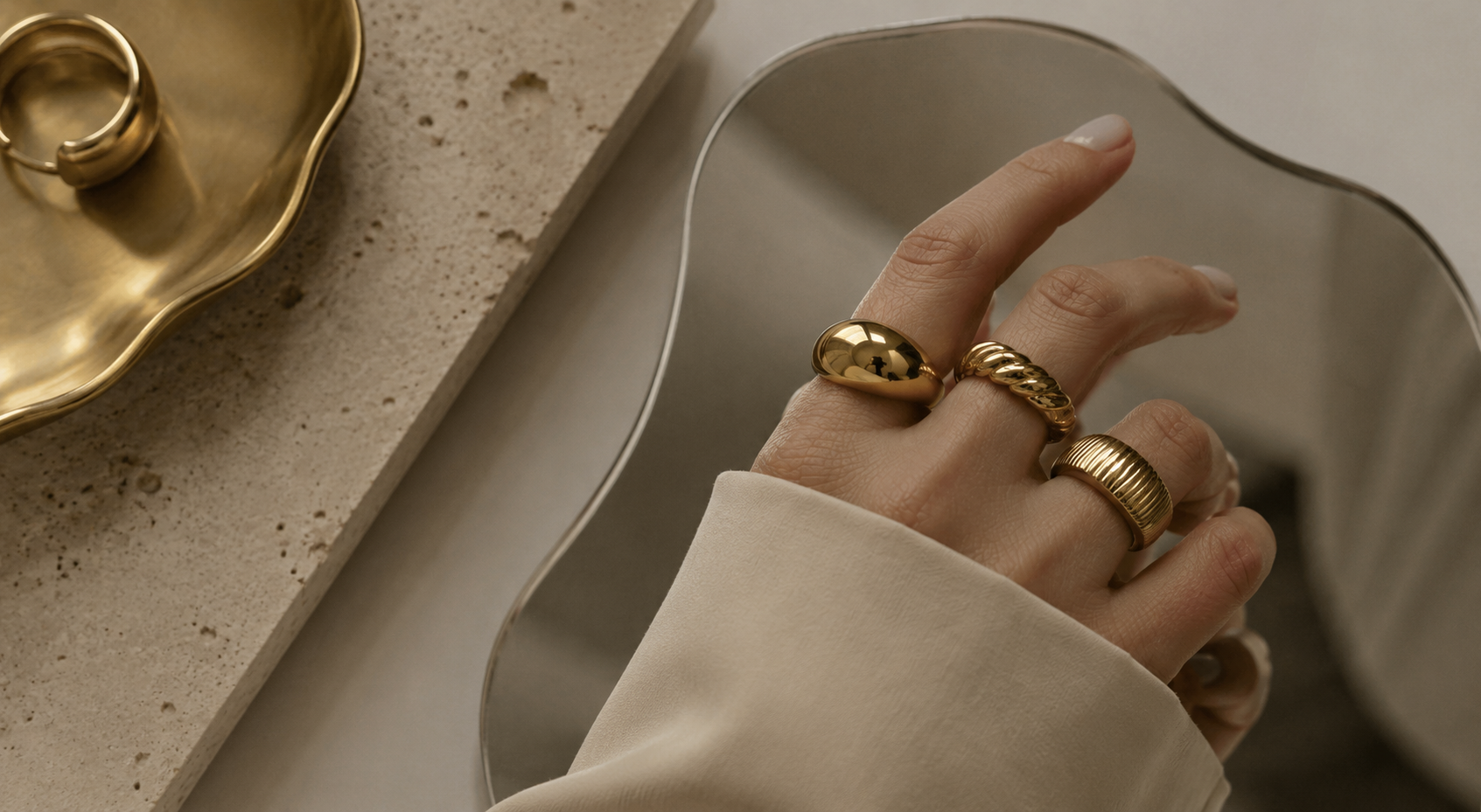 Minimal editorial close-up of a hand wearing chunky gold rings beside a sculptural metallic tray and textured stone surface in warm neutral lighting.
