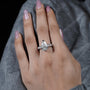 Hand wearing a diamond ring with pink nail polish on a gray textured background

