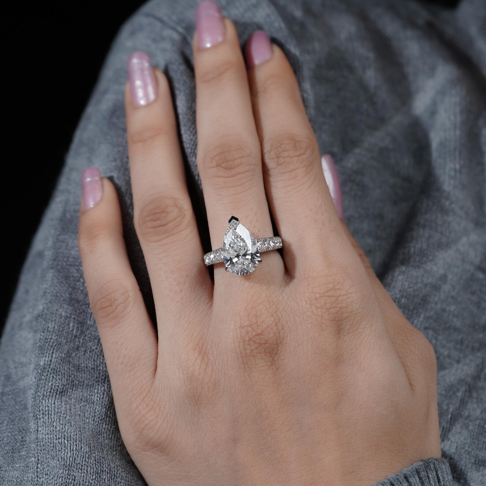Hand wearing a diamond ring with pink nail polish on a gray textured background

