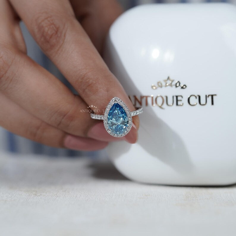 Pear-shaped blue gemstone ring held by a hand with 'AntiqueCut' branding in the background.