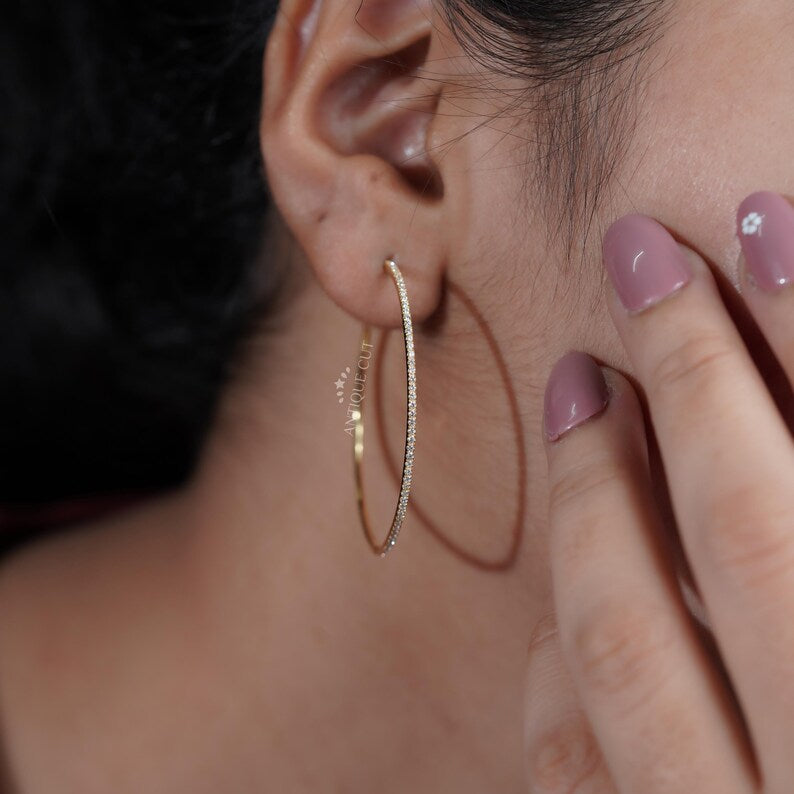 Gold hoop earrings held by a hand with a blurred 'Antique Cut' container in the background.