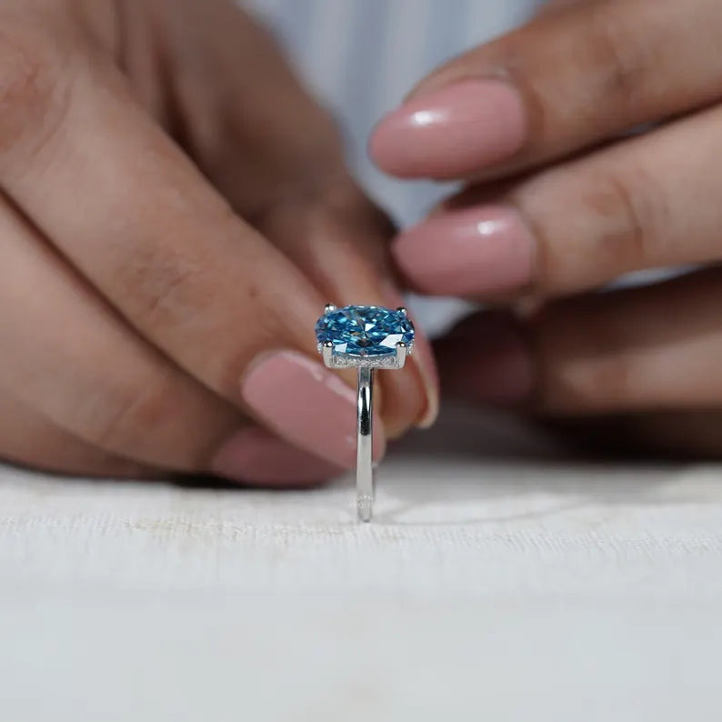 Blue gemstone ring held by a hand with 'AntiqueCut' branding in the background.
