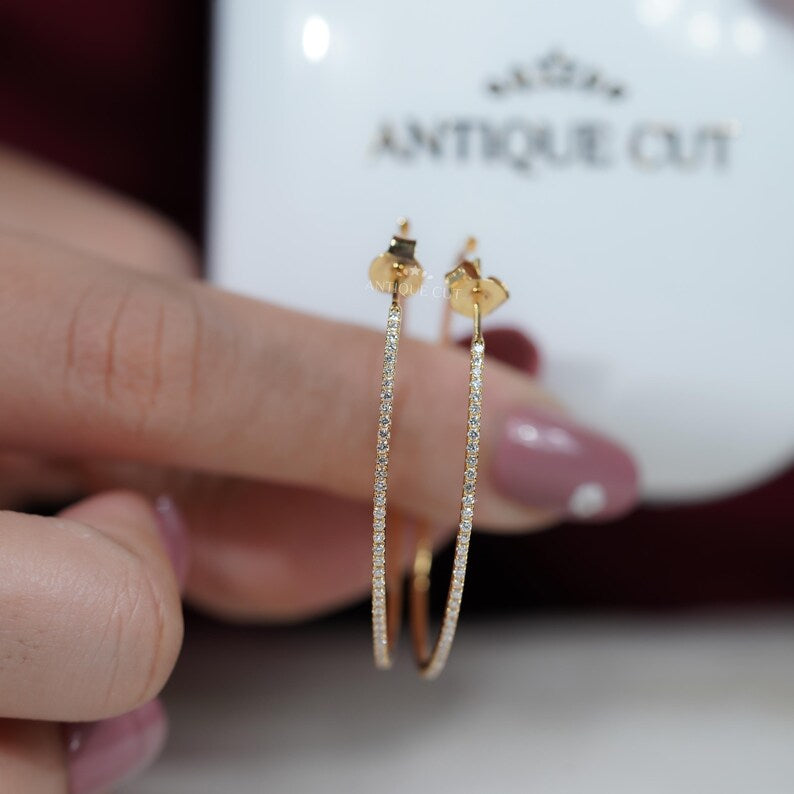 Gold hoop earrings held by a hand with a blurred 'Antique Cut' container in the background.