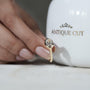 Gold ring with a diamond held by a hand in front of an 'Antique Cut' branded container.


