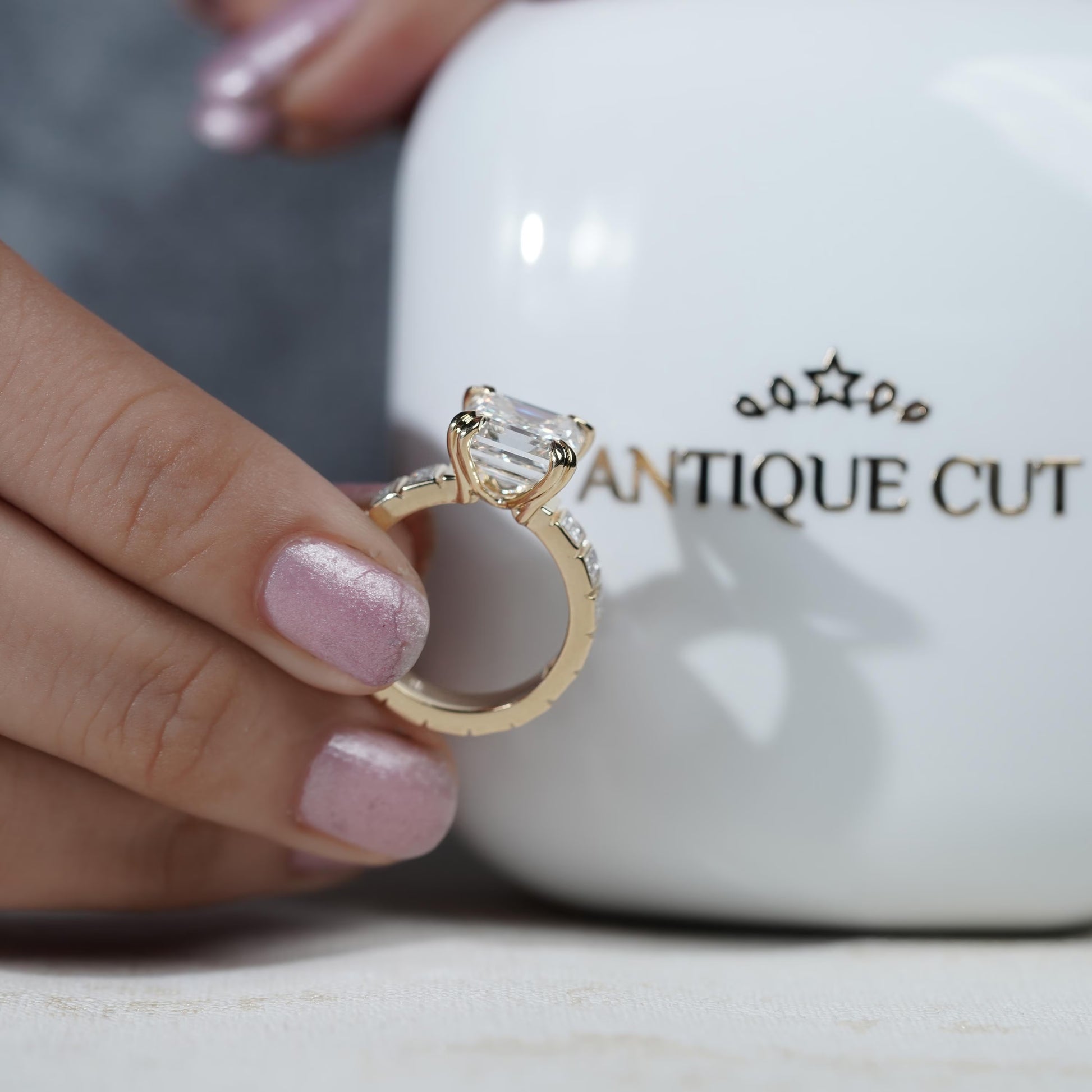 Gold ring with a diamond held by a hand in front of a 'Antiquecut' branded ring box 

