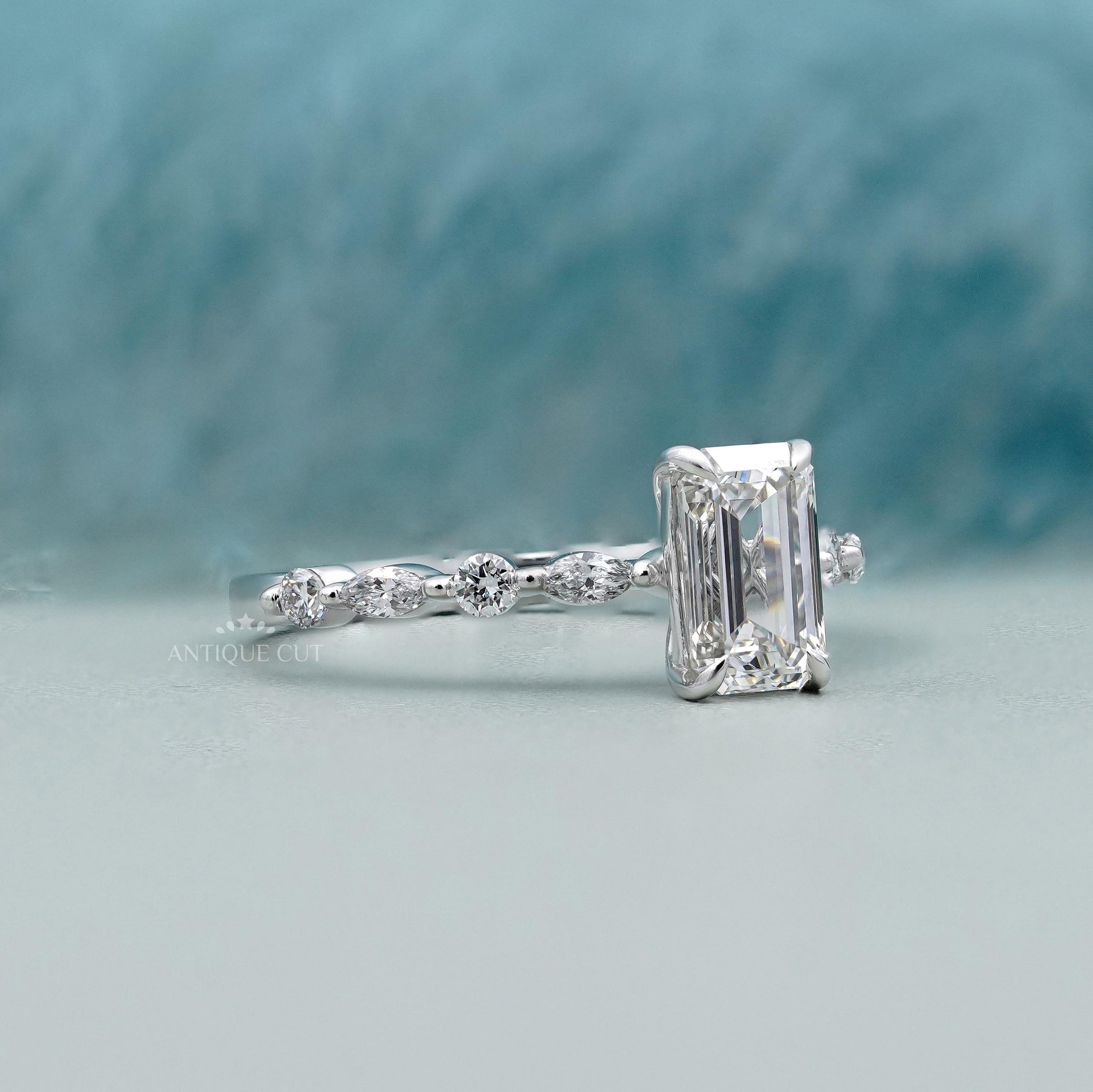 Diamond ring with emerald-cut center stone on a light blue background

