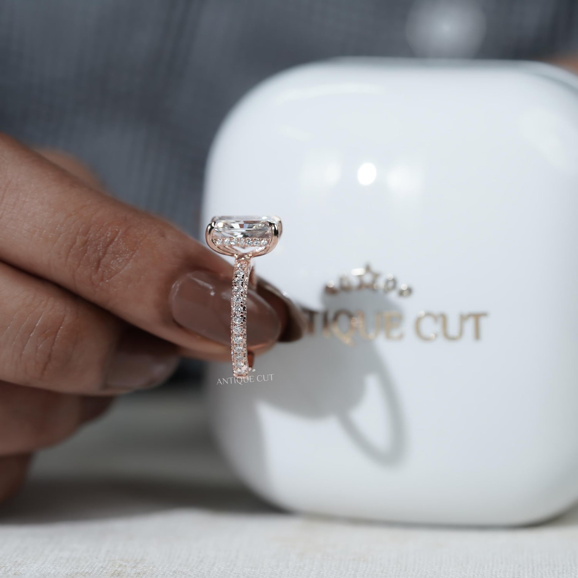 Diamond ring held by a hand with a blurred 'antiquecut' container in the background
