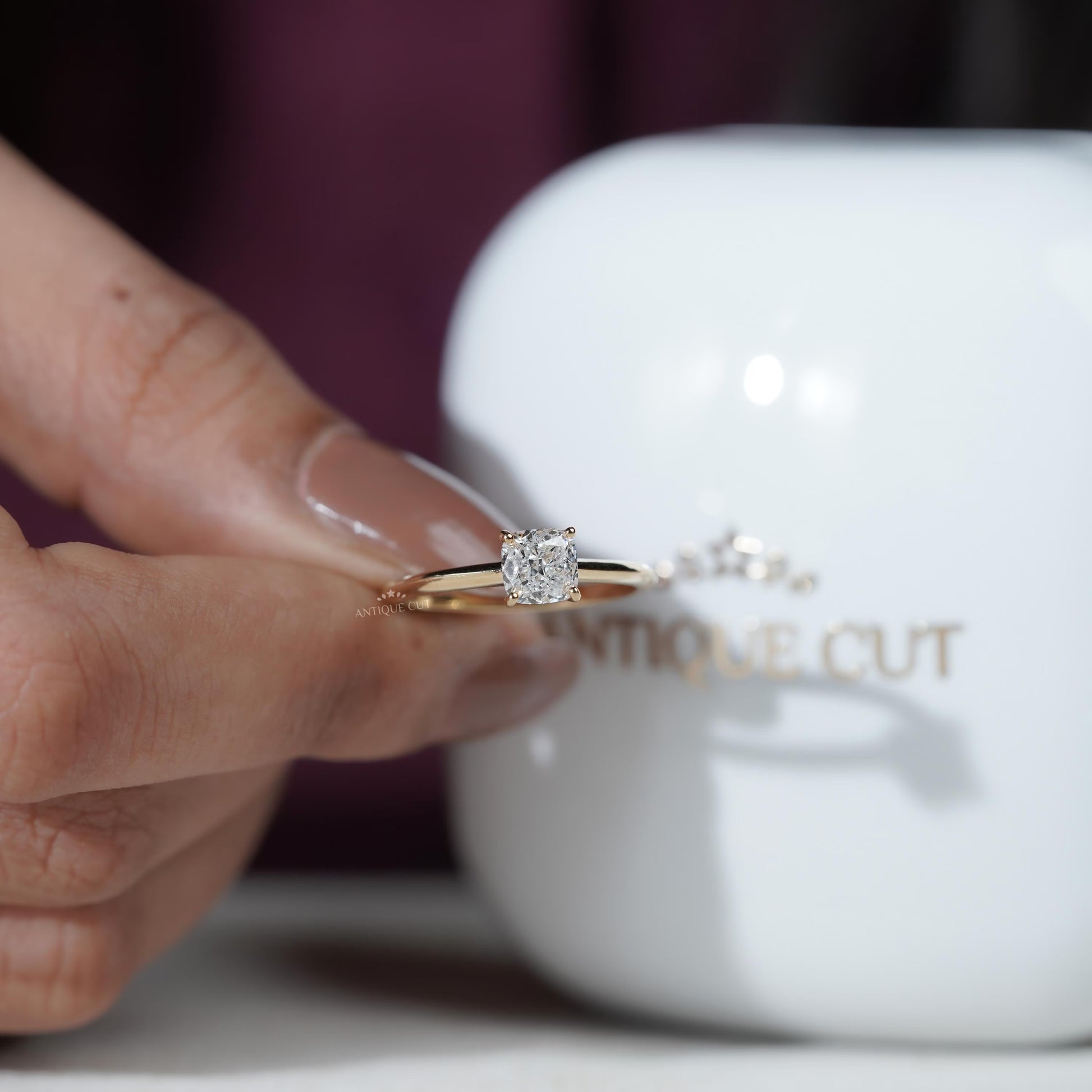 Gold ring with a diamond held by a hand in front of an Antique Cut