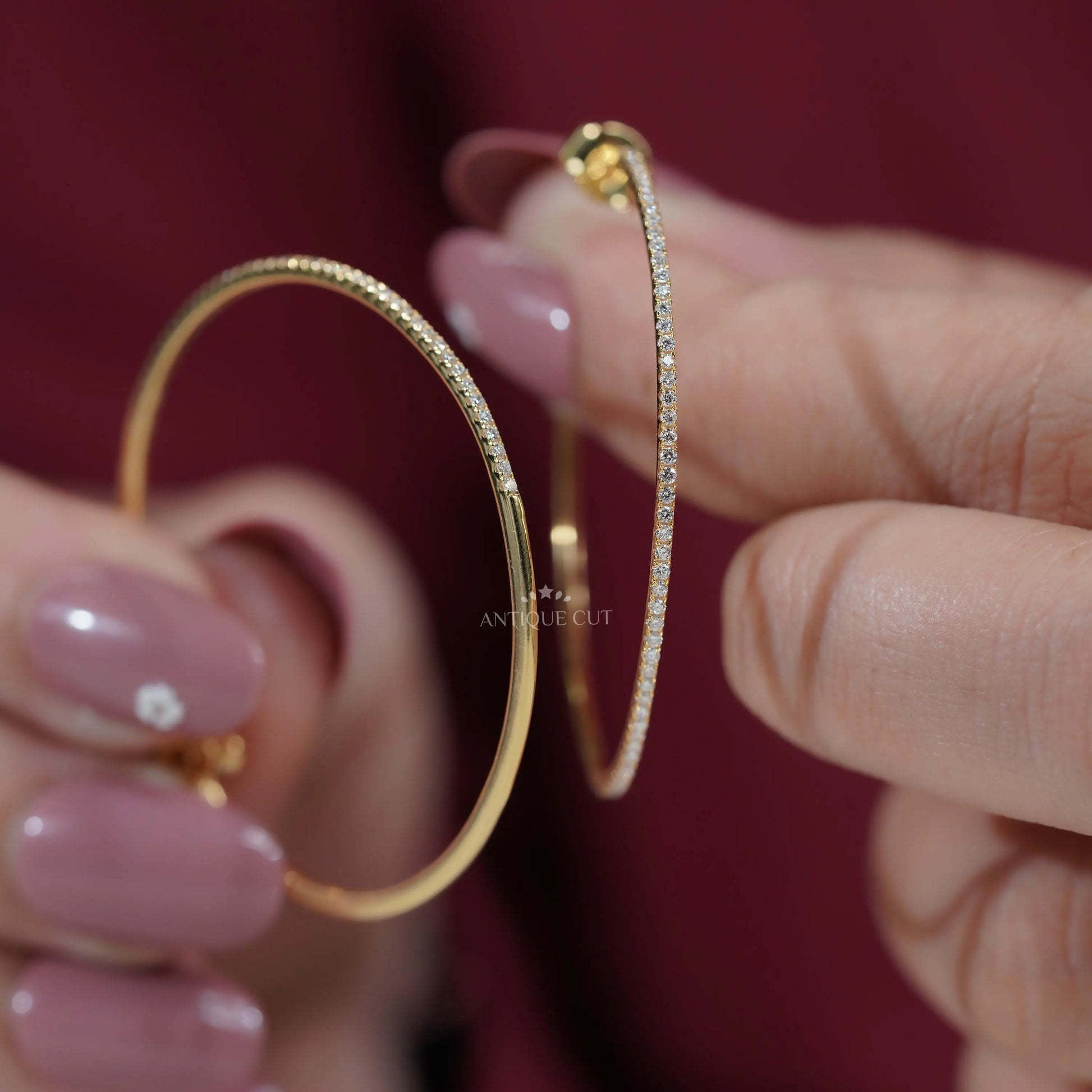 Gold hoop earrings held by a hand with a blurred 'Antique Cut' container in the background.