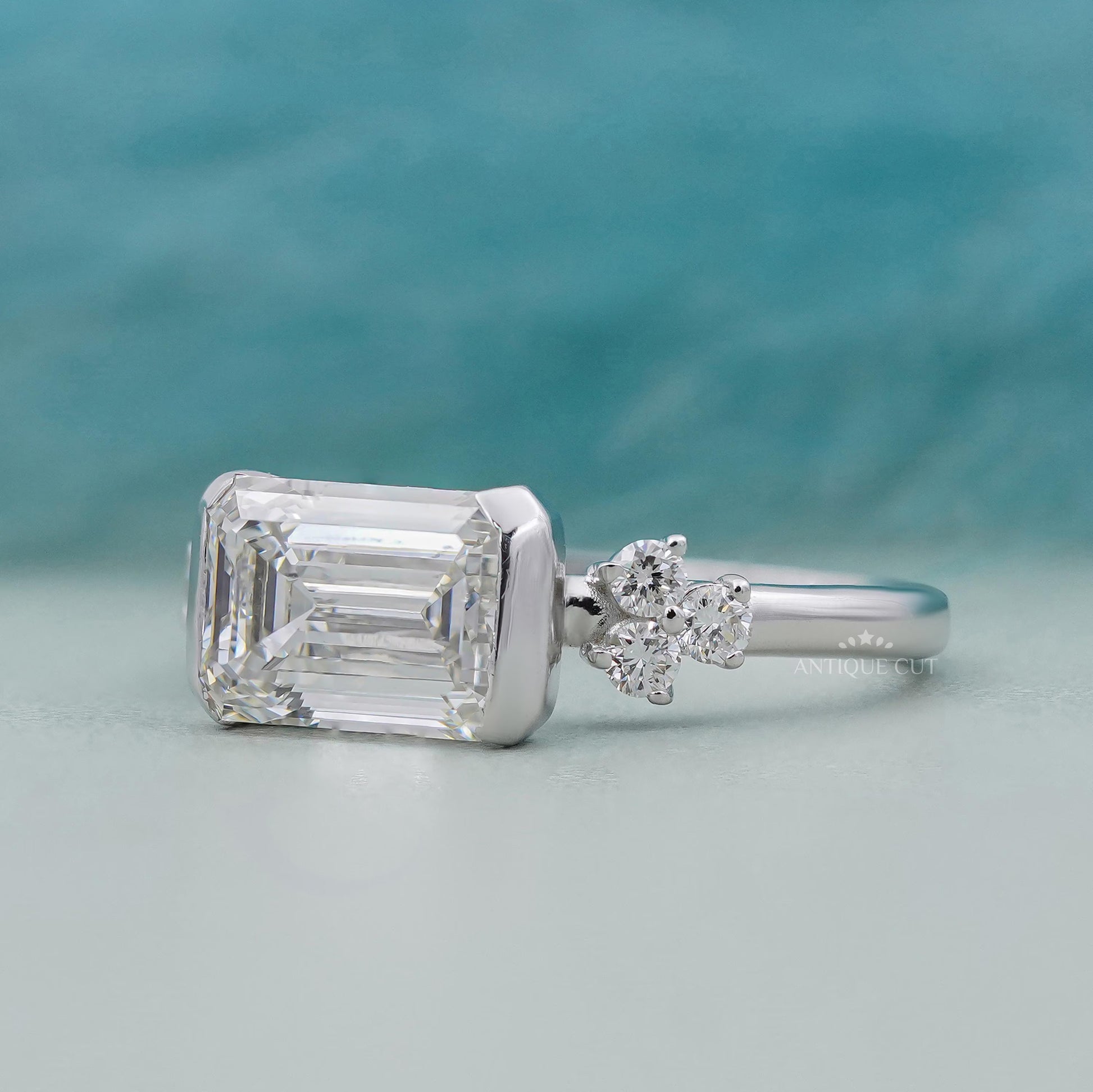 Silver ring with emerald-cut diamond and smaller diamond on a teal background


